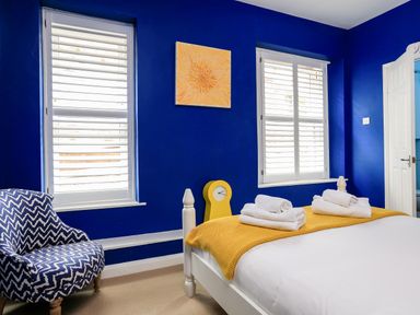 A bedroom with blue walls white framed windows a bed with white bedding and yellow blanket a patterned blue and white chair a yellow clock and an orange painting at Armorel House in Dartmouth