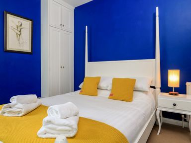 A bedroom with a white bed with yellow pillows and blanket beside a white nightstand with a lamp and blue walls at Armorel House in Dartmouth