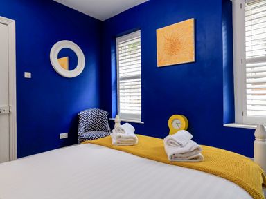 A bedroom with a bed, folded towels, a blue and white patterned chair, two windows with white shutters, a round white mirror, a yellow clock, and a yellow artwork on blue walls at Armorel House in Dartmouth