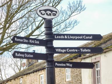 A signpost with directions to Roman Site Kirk Sink Leeds Liverpool Canal Village Centre Toilets Railway Station and Pennine Way near stone buildings