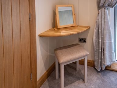 A corner with a wooden floating shelf holding a mirror a fabric stool on carpet next to a window with curtains at Otter Lodge in Gargrave