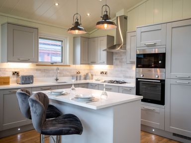 A kitchen with grey cabinets an island with bar stools and hanging lights at Otter Lodge in Gargrave