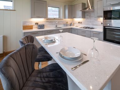 A kitchen with gray cabinets a white countertop island with place settings and upholstered chairs at Otter Lodge in Gargrave