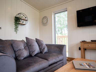 A living room with a gray sofa pillows a wall clock a wall shelf with plants a wooden table and a wall-mounted TV at Otter Lodge in Gargrave