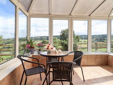 A sunroom with a round table and three chairs with a view of green fields and hills at The Stables At Cae Gwyn in Ruthin