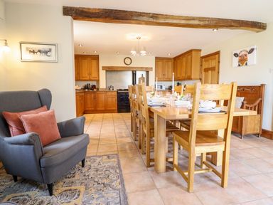 A dining area with a wooden table and chairs set for a meal next to a kitchen with wooden cabinets and a gray armchair at The Stables At Cae Gwyn in Ruthin