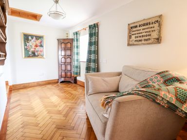 A room with a beige sofa covered with a checkered blanket a wooden corner cabinet patterned curtains and framed art at The Stables At Cae Gwyn in Ruthin