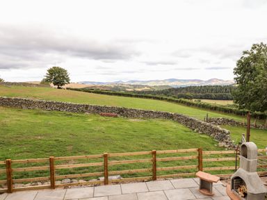 A patio with a stone fireplace and wooden bench overlooking a fenced grassy field and stone wall with hills in the background at The Stables At Cae Gwyn in Ruthin