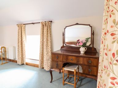 A room with a wooden dressing table with a mirror flowers in a jug and a stool next to a window with floral curtains at The Stables At Cae Gwyn in Ruthin