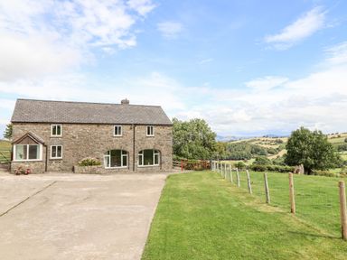 A stone house with three windows and a driveway beside a grassy fenced yard with trees and hills in the background at The Stables At Cae Gwyn in Ruthin