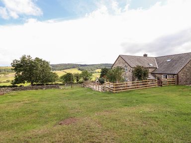 A stone house with wooden fencing and green lawn with trees and rolling hills in the background at The Stables At Cae Gwyn in Ruthin