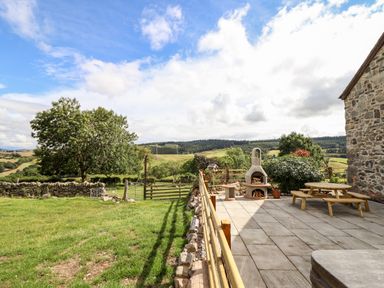 A patio with outdoor fireplace and wooden picnic bench next to a stone wall overlooking green fields and trees at The Stables At Cae Gwyn in Ruthin