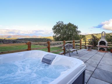 An outdoor patio with a hot tub bench stone grill and wooden fence overlooking trees and hills at The Stables At Cae Gwyn in Ruthin
