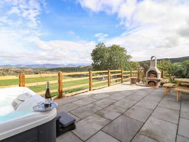 An outdoor patio with a hot tub and a bottle of wine on a tray and a barbecue oven with picnic tables and countryside views at The Stables At Cae Gwyn in Ruthin