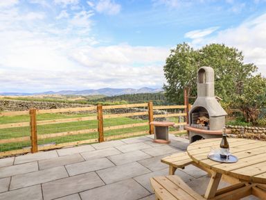 An outdoor patio with a wooden picnic table holding a bottle and glasses next to a stone fireplace and a wooden fence overlooking a grassy field and hills at The Stables At Cae Gwyn in Ruthin