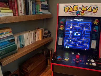 A pac man arcade machine next to wooden bookshelves filled with books and a wooden chest on the floor at The Stables At Cae Gwyn in Ruthin