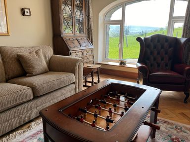A living room with a foosball table sofa armchair cabinet and large window showing green fields at The Stables At Cae Gwyn in Ruthin