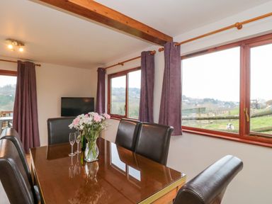 A dining room with a wooden table, leather chairs, flowers in a vase, and windows showing an outdoor view at The Willow Lodge near Lyme Regis