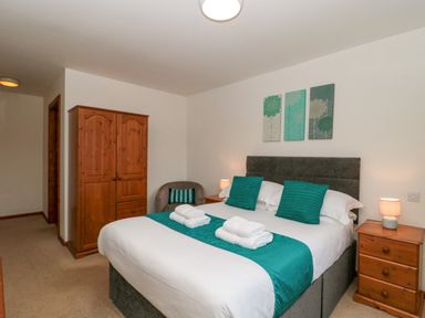 A bedroom with a double bed with teal cushions and runner bedside tables with lamps a wooden wardrobe and a chair at The Willow Lodge near Lyme Regis