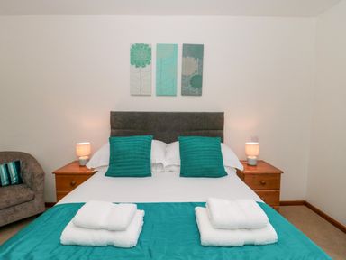 A bedroom with a bed with teal pillows and blanket two wooden bedside tables with lamps and a gray armchair near The Willow Lodge near Lyme Regis