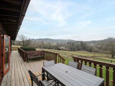 A wooden outdoor deck with a table and chairs and a bench overlooking a rural landscape at The Willow Lodge near Lyme Regis