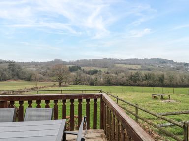 A wooden deck with table and chairs overlooking a fenced grassy field and distant hills at The Willow Lodge near Lyme Regis