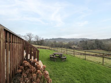 A garden with a wooden picnic table and a wooden railing next to grass and hills at The Willow Lodge near Lyme Regis