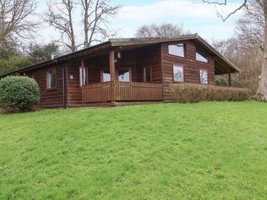 A wooden cabin with a porch and multiple windows on a grassy slope near trees at The Oak Lodge near Lyme Regis