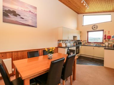A dining area with a wooden table and six black chairs next to a kitchen with white cabinets and a window at The Oak Lodge near Lyme Regis