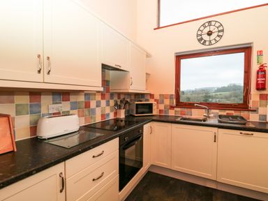 A kitchen with cream cabinets a black countertop a toaster a microwave a stove a sink a wall clock and a window showing a countryside view at The Oak Lodge near Lyme Regis