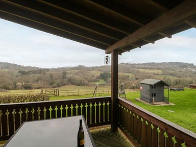 A covered wooden porch with a glass table and a bottle overlooking a grassy yard with a playhouse and swing set at The Oak Lodge near Lyme Regis