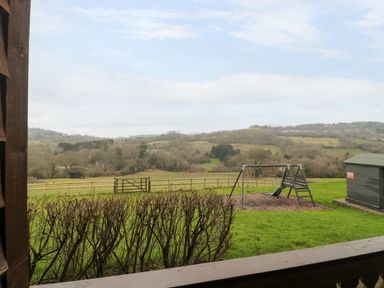 A view of a playground with swings and a wooden shed in a grassy area with hills and trees in the background at The Oak Lodge near Lyme Regis