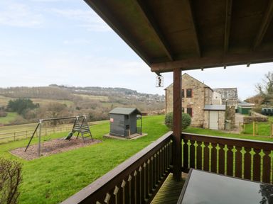 A covered wooden porch overlooking a grassy yard with a swing set and a small playhouse near a stone building at The Oak Lodge near Lyme Regis