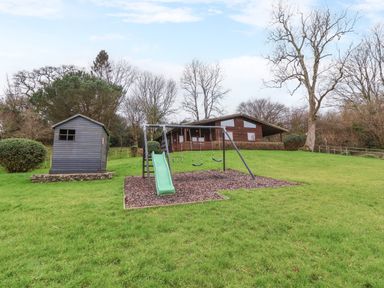 A playground with swings and a slide near a small shed and a wooden house in a grassy area at The Oak Lodge near Lyme Regis