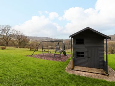 A playhouse and swing set in a grassy yard with trees and hills in the background at The Oak Lodge near Lyme Regis
