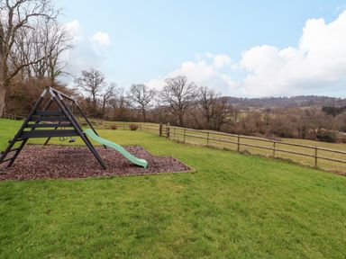 An outdoor playground with a slide and swings on a grassy field with trees and a wooden fence at The Oak Lodge near Lyme Regis