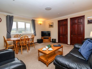 A living room with a wooden dining table and chairs a television on a wooden stand and black leather sofas at The Hayloft Cottage Apartment near Lyme Regis