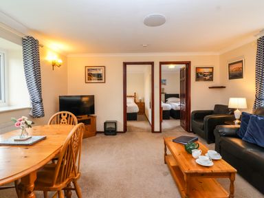 A living room with wooden dining table and chairs black sofa and armchair a coffee table with teacups television and two doorways showing bedrooms at The Hayloft Cottage Apartment near Lyme Regis