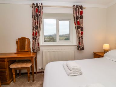 A bedroom with a bed with towels folded on it a wooden dressing table with a mirror a cushioned stool patterned curtains and a window showing a rural view at The Hayloft Cottage Apartment near Lyme Regis