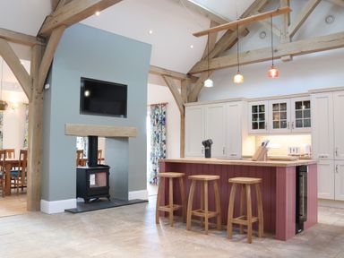A kitchen with an island and three stools under pendant lights and a stove below a mounted television at The Anchorage @ Nables Farm in Upper Seagry