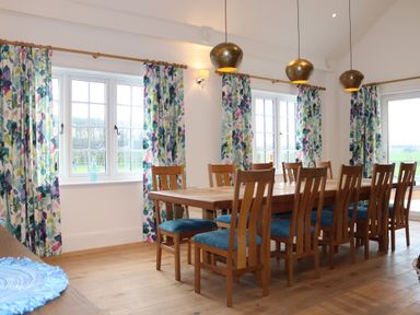 A dining room with a long wooden table and ten wooden chairs with blue cushions and floral curtains at The Anchorage @ Nables Farm in Upper Seagry