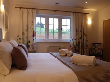 A bedroom with a bed pillows folded towels floral curtains and a chair at The Anchorage @ Nables Farm in Upper Seagry