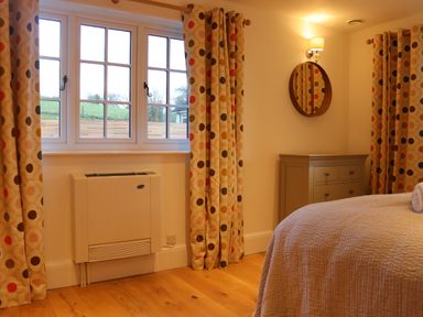 A bedroom with a bed a chest of drawers a wall mirror and a window with patterned curtains at The Anchorage @ Nables Farm in Upper Seagry
