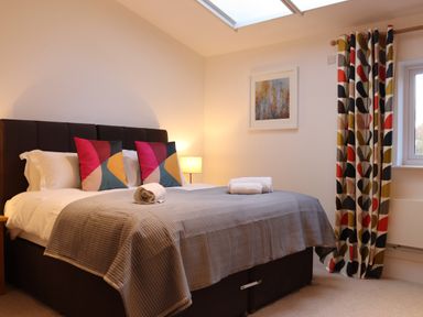 A bedroom with a double bed with colorful pillows bedside lamp window with patterned curtains and a framed picture on the wall at The Anchorage @ Nables Farm in Upper Seagry