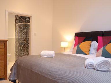 A bedroom with a bed covered in a gray bedspread and rolled towels with colorful pillows and a lamp next to an ensuite bathroom with patterned tiles at The Anchorage @ Nables Farm in Upper Seagry