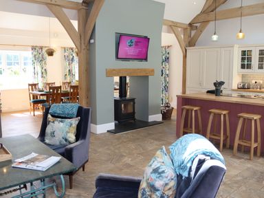 An open plan living room with armchairs and cushions a kitchen with stools and a dining area with a wooden table and chairs at The Anchorage @ Nables Farm in Upper Seagry