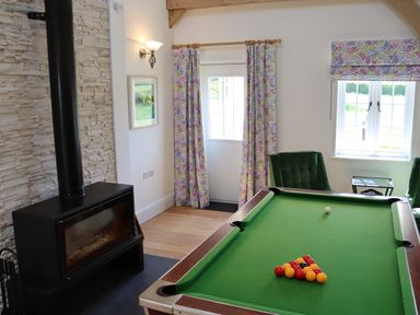 A room with a pool table a black wood stove two green chairs and windows with patterned curtains at The Anchorage @ Nables Farm in Upper Seagry