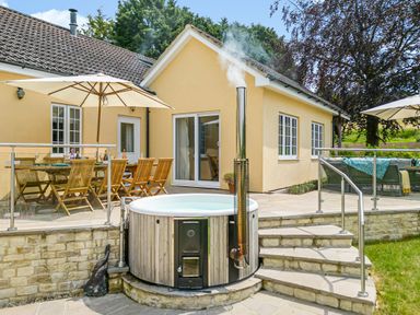 An outdoor patio area with wooden dining chairs and table under umbrellas and a round hot tub at The Anchorage @ Nables Farm in Upper Seagry