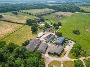 An aerial view of a farm complex with multiple buildings surrounded by green fields and trees at The Anchorage @ Nables Farm in Upper Seagry
