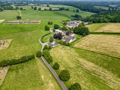 An aerial view of a rural farm complex with multiple buildings surrounded by large fields and trees at The Anchorage @ Nables Farm in Upper Seagry